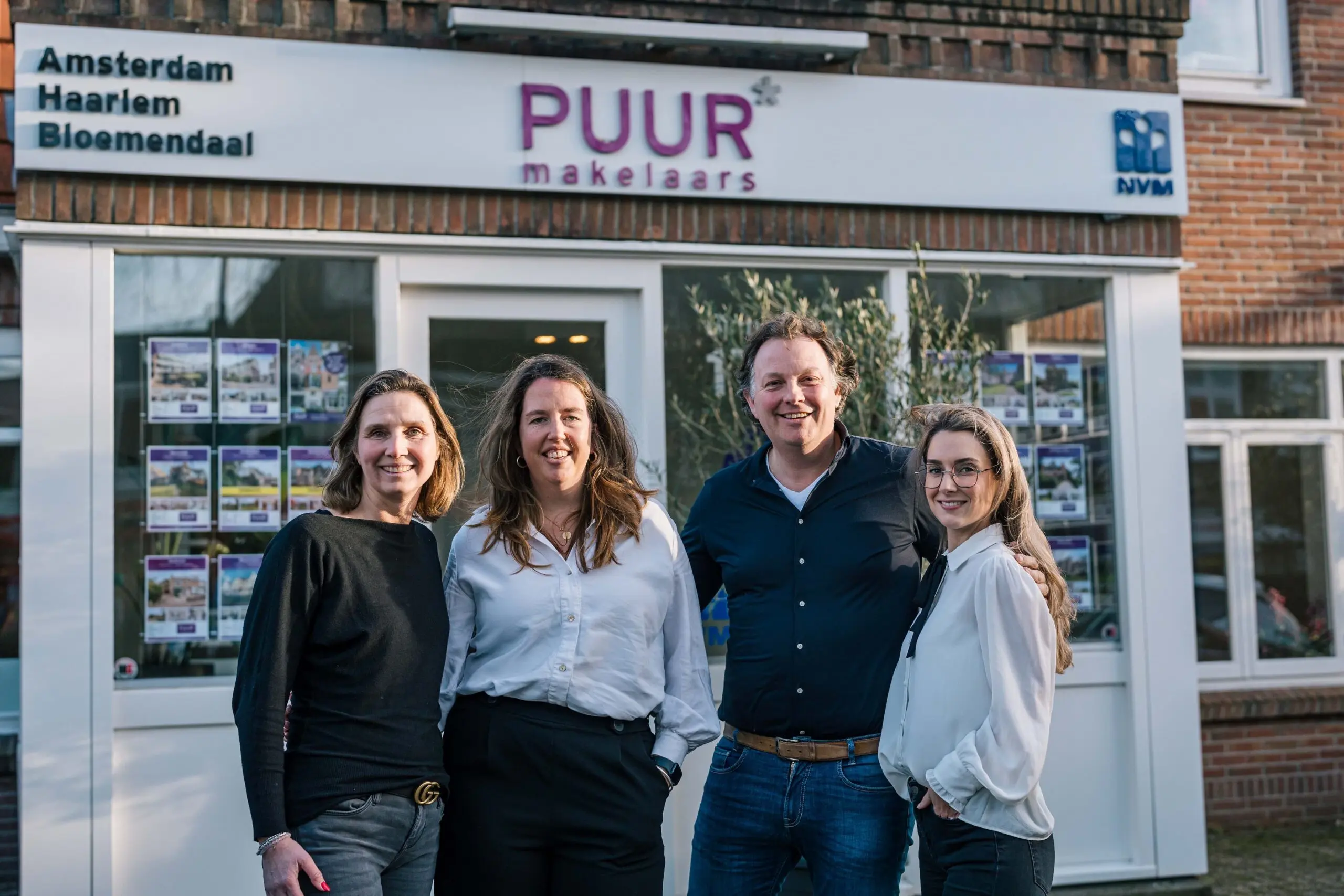 A group of four professionals standing in front of the PUUR makelaars real estate office, showcasing property listings in the window.