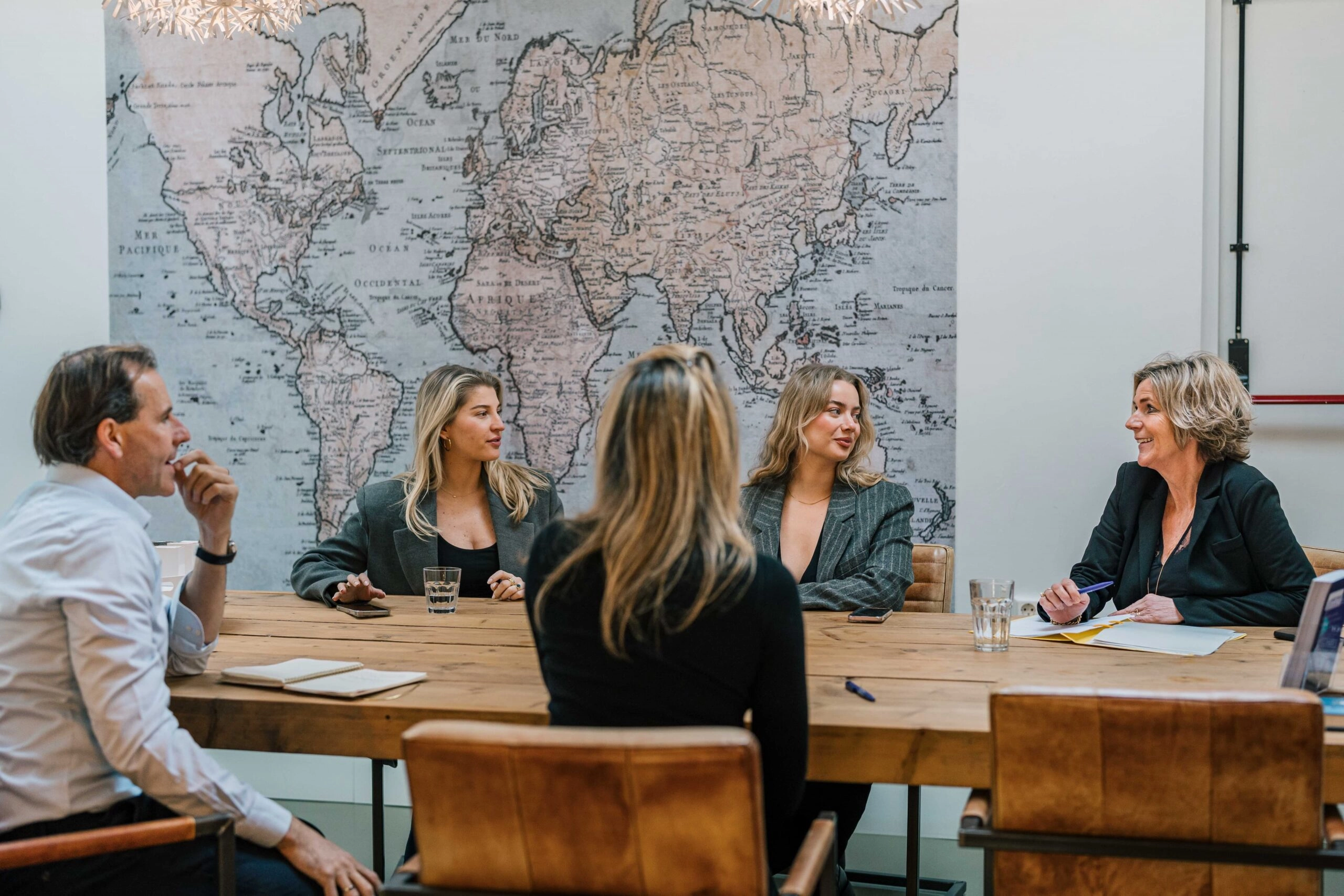 Five people, three women and two men, are engaged in a business meeting around a wooden table. They are seated in a modern conference room with a large vintage world map on the wall in the background. The group appears to be in a collaborative discussion, with notepads and glasses of water on the table.