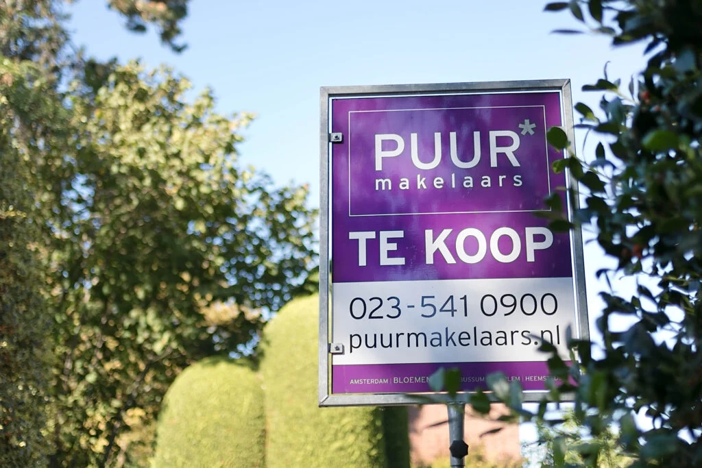 Een levendig paars 'Te Koop' bord van PUUR makelaars te midden van weelderig groen, wat wijst op een veelbelovende vastgoedkans in een serene omgeving.