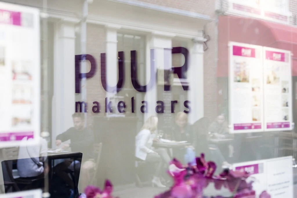 De levendige weerspiegeling van de 'PUUR makelaars' bewegwijzering overlapt het levendige, maar vage tafereel van een bruisend café-interieur.