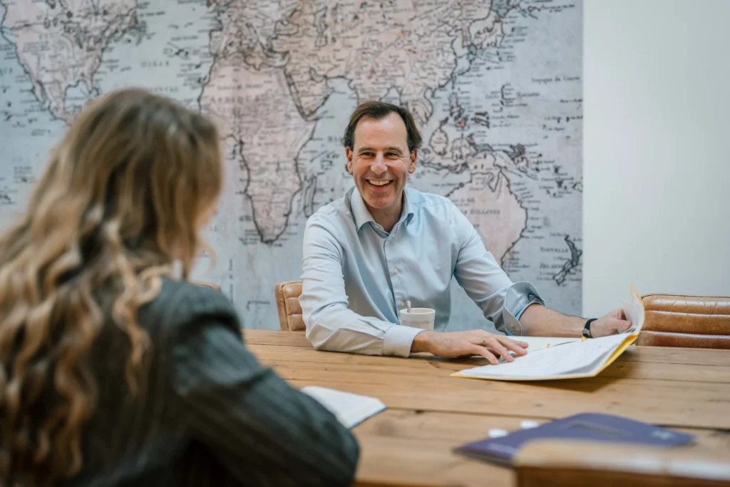 Een man in een lichtblauw shirt lacht en praat met een vrouw met lang krullend haar aan de andere kant van een houten tafel, met een wereldkaart op de muur achter hen.