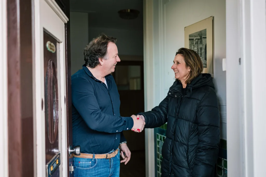 Een man in een blauw shirt en een vrouw in een zwarte jas schudden elkaar de hand bij een deuropening, beide glimlachend.