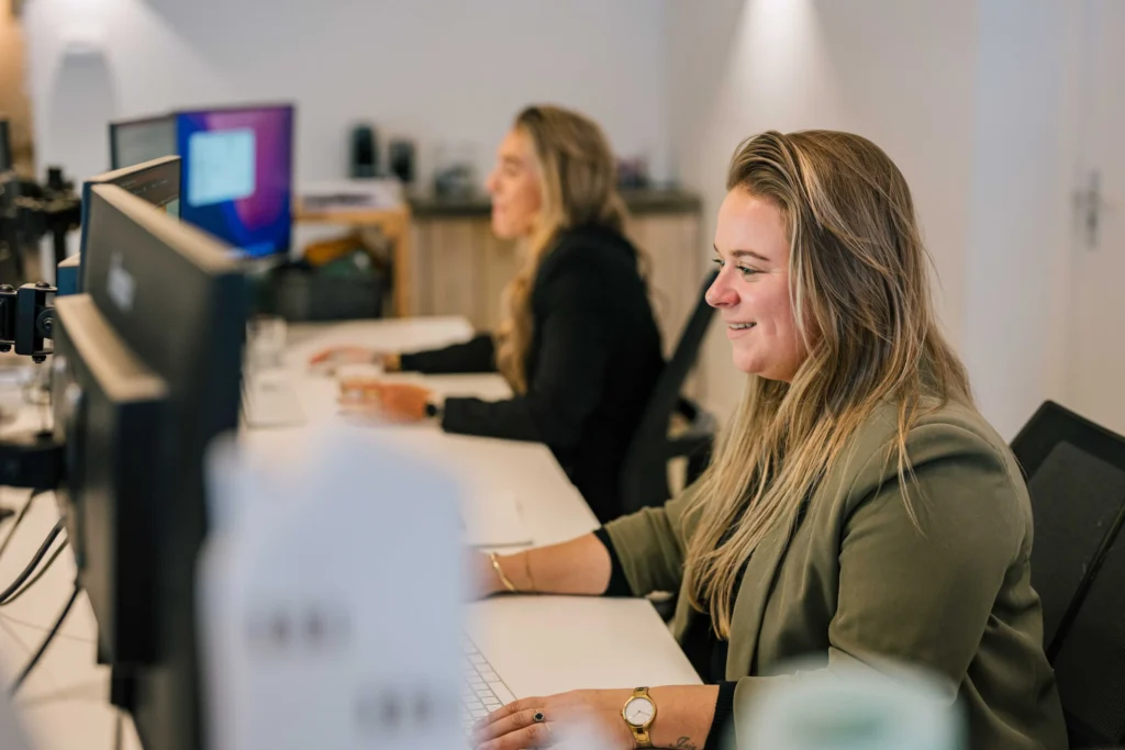 Twee vrouwen werken aan bureaus in een kantoor, gefocust op hun computerschermen.