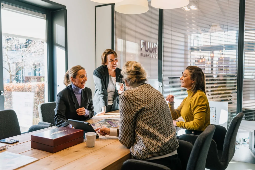 Vier vrouwen hebben een levendig gesprek rond een houten tafel in een kantoor met grote ramen en een "PUUR makelaars" bord op de achtergrond.