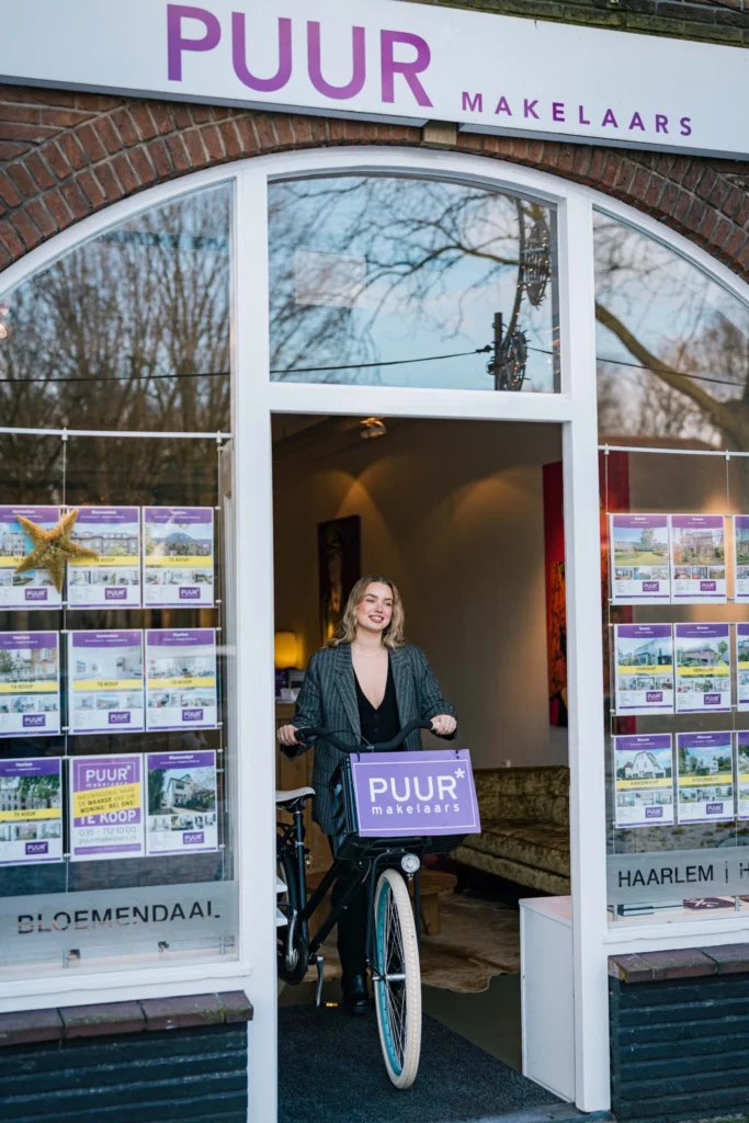 Een vrouw staat met een fiets voor een "PUUR makelaars" makelaarskantoor, met woningaanbiedingen in de ramen.