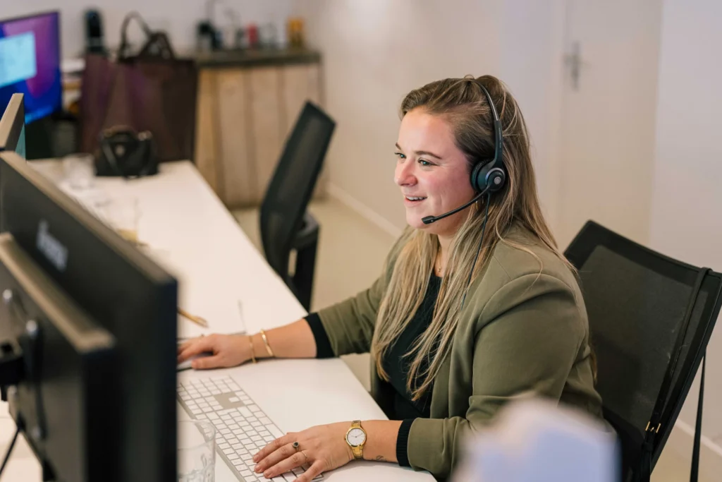 Een vrouw met een headset zit aan een bureau en typt op een toetsenbord terwijl ze naar een computerscherm kijkt.