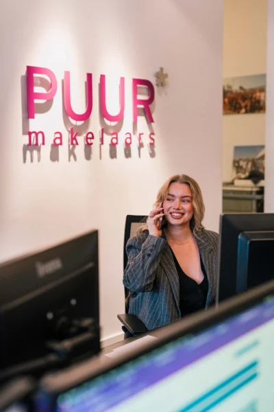 Een vrouw zit aan een bureau, lacht en praat aan de telefoon, met het bord "PUUR makelaars" op de muur achter haar.